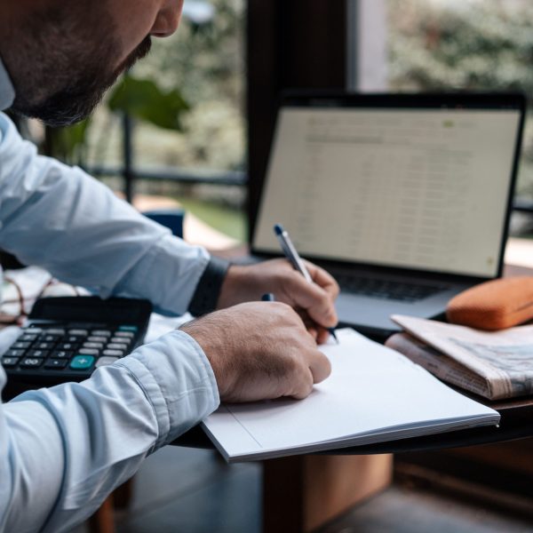 A man writing in a notebook while using a laptop and calculator at a desk.